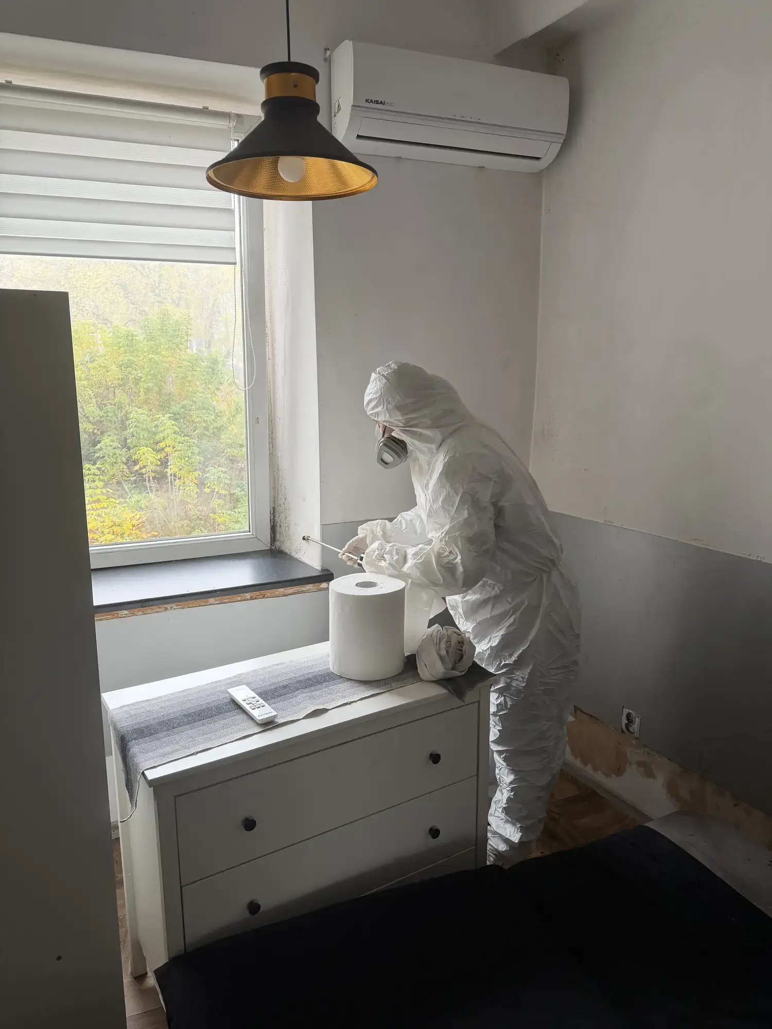 Pracownik w kombinezonie z kapturem kieruje lancę na narożnik wnęki okiennej przy parapecie